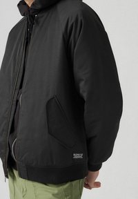 Svart bomberjacka med dragkedja fram, sidofickor, ribbade mudd och fåll. Slät väv och något vadderad för värme.