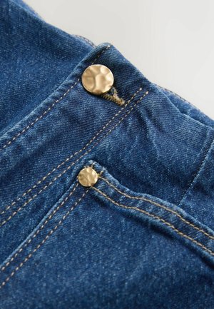 Material din denim albastru închis cu închizători metalice aurii, având cusături contrastante bej de-a lungul marginilor și deschiderilor buzunarelor.