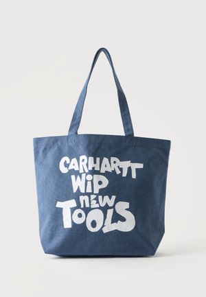 Blå lærreds tote taske med lange håndtag og hvid fed tekst med "Carhartt WIP new Tools" på forsiden.