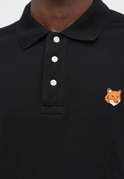 Detailní záběr osoby v černém polokošili se třemi bílými knoflíky a vyšitou pomerančovou hlavou lišky na levé části hrudi.