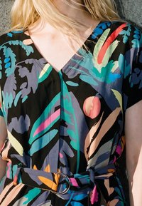 Personne blonde portant une robe colorée à manches courtes avec des motifs abstraits de feuilles et de formes et une taille ceinturée sur fond gris.