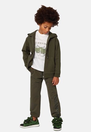 Olivgrüner Kapuzenpullover mit Reißverschluss und Kordelzügen, weißes Grafik-T-Shirt, passende Jogginghose und lebendige grüne Sneakers mit strukturierten Akzenten.