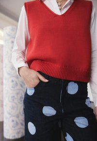 Piros kötött ujjatlan póló V-nyakkal és bordás szegéllyel, sötét farmernadrággal párosítva, amelyen nagy világoskék pöttyök találhatók. Kéz a zsebben.