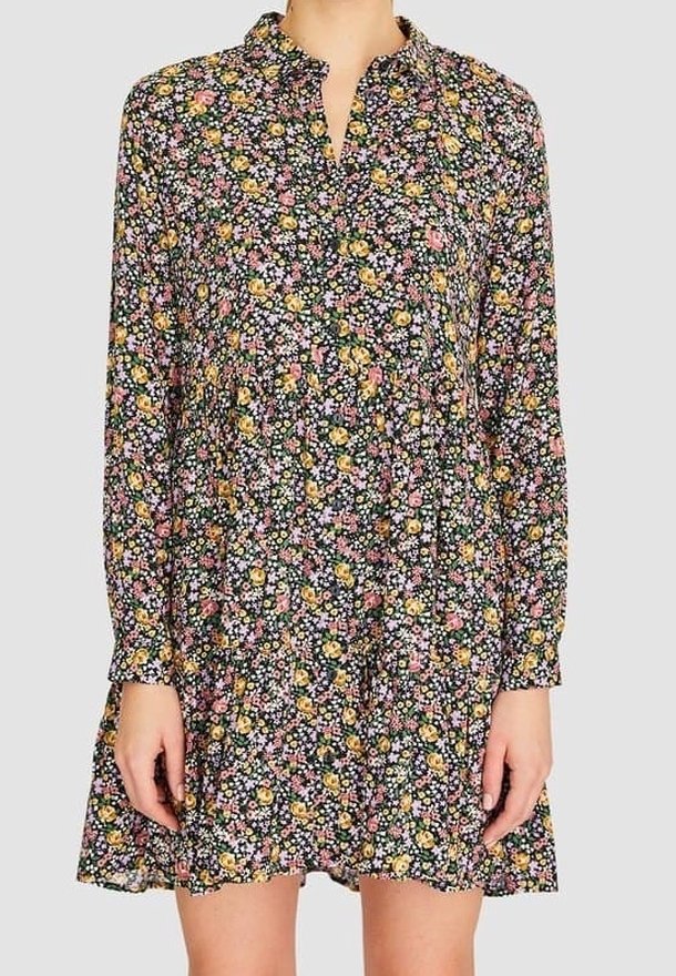 Femme portant une robe fleurie à manches longues, au-dessus du genou, avec un col à volants et un mélange de fleurs jaunes, roses et blanches sur fond sombre.