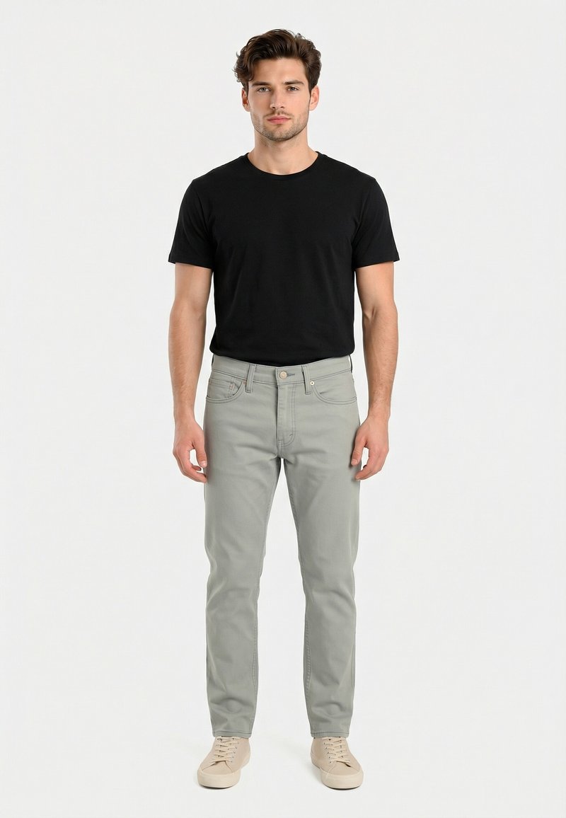 Hombre de pie vistiendo una camiseta negra de manga corta, vaqueros gris claro y zapatillas beige sobre un fondo blanco liso.