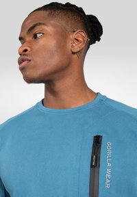 Blauwe sweatshirt met ronde hals van textuurstof. Voorzien van een ritszak op de borst en een "GORILLA WEAR" logo ernaast.