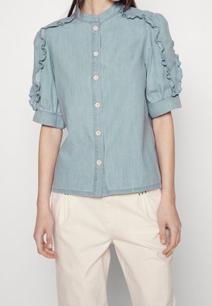 Femme portant un chemisier bleu clair à boutons avec des manches courtes volantées et un pantalon beige taille haute, debout devant un fond uni.