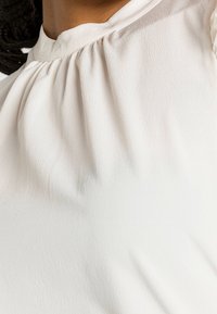 Blouse blanche légère avec tissu froncé au niveau du cou, surface légèrement texturée et design doux et fluide.