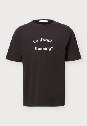 Мъж представя тъмна тениска с надпис „California Running“ отпред на изчистен светъл фон.
