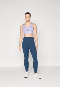 Svetlo vijolični športni nedrček z napisom "Nike Pro", v kombinaciji z mornarsko modrimi leginami, ki imajo stranske žepke z zadrgo in visok pas. Bele športne copate.