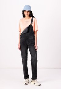 Zwarte denim overalls met een zijzak, gedragen over een lichtroze graphic t-shirt. De outfit wordt aangevuld met een blauwe bucket hat en sneakers.