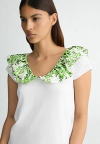 Haut blanc à manches courtes avec un col froncé à motifs verts et blancs. Le tissu est lisse, avec une coupe décontractée pour le confort.