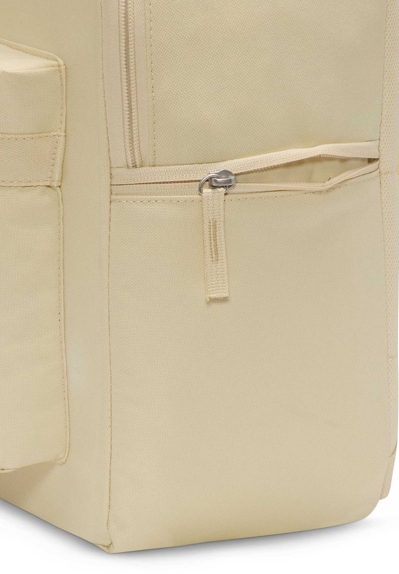 Mochila de tela beis con una superficie texturizada, que presenta un bolsillo frontal con cremallera y un bolsillo de malla lateral para almacenamiento adicional.
