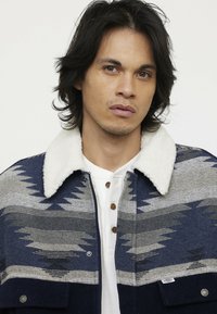 Wollmischung-Jacke mit einem blauen und grauen geometrischen Muster, weichem weißen Kragen, vorderen Knöpfen und einer unteren Tasche mit einem Logo.