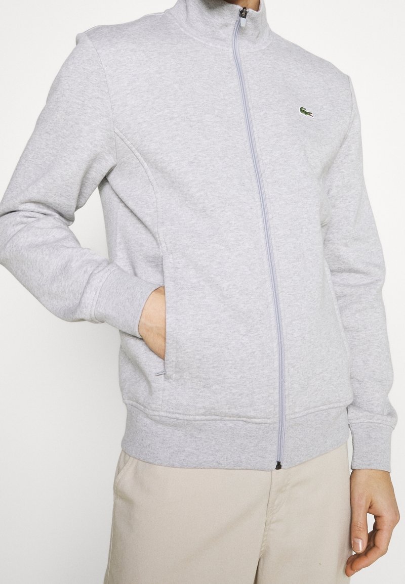 Ljusgrå zip-tröja i mjukt material. Har en hög krage, ribbade ärmslut och en liten broderad logotyp på bröstet.