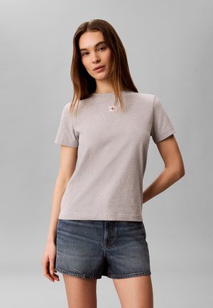 Jonge vrouw met lang bruin haar die een lichtgrijs T-shirt met korte mouwen en donkere denim shorts draagt, staand tegen een effen grijze achtergrond.