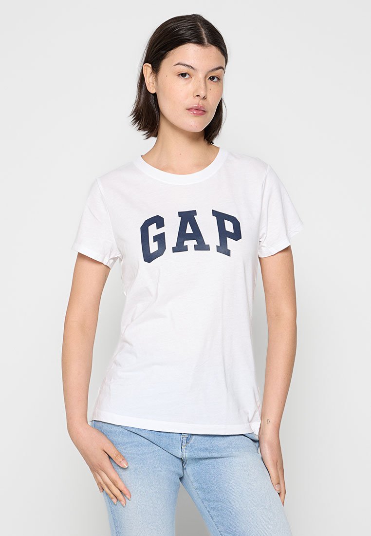 Camiseta de algodón blanca con un logo azul de "GAP", escote redondo, mangas cortas y un diseño ajustado, combinada con pantalones vaqueros de un azul claro.