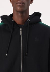 Svart hettegenser med sølvglidelås, med et grønt gradientdesign på skuldrene og en logo brodert på brystet.