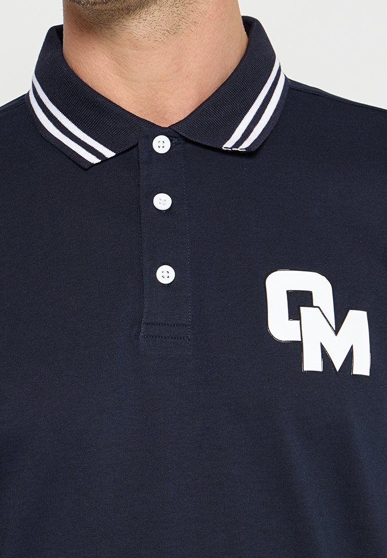 Granatowa koszulka polo z paskowanym kołnierzykiem w białym i granatowym kolorze, trzema białymi guzikami oraz białym wzorem logotypu po lewej stronie klatki piersiowej.