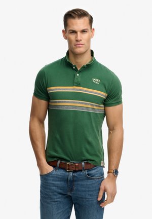 Jonge man met kort bruin haar, gekleed in een groen poloshirt met gele en grijze strepen, blauwe spijkerbroek, bruine riem en polshorloge.