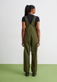 Olive green overalls with a V-shaped back, wide legs, and two back pockets, paired with a fitted black t-shirt and chunky black boots.