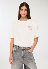 Młoda kobieta z długimi ciemnymi włosami, ubrana w białą koszulkę z różowym napisem oraz wyblakłe szare dżinsy, z jedną ręką w kieszeni, na neutralnym tle.