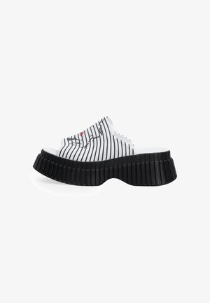 Mules blanches avec des rayures verticales noires, bout ouvert arrondi et semelle épaisse en caoutchouc noir texturé avec des rainures. Détail accentué en rouge.
