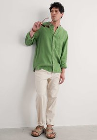 Camicia button-up in lino verde, pantaloni beige e sandali marroni. La camicia presenta una tasca sul petto e una vestibilità comoda con maniche arrotolate.