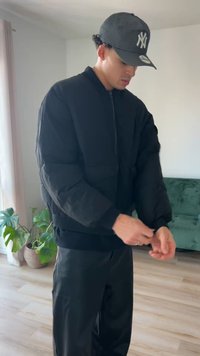 Veste matelassée noire avec fermeture éclair devant, poignets et ourlet côtelés. Portée avec un pantalon noir assorti. En arrière-plan, un canapé vert et des plantes.