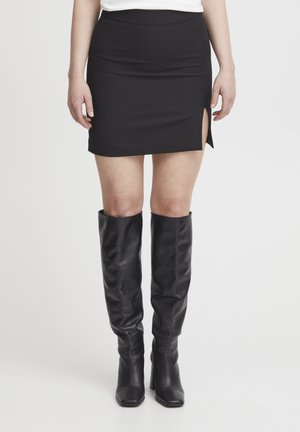 Schwarzer Minirock mit seitlichem Schlitz, aus einem glatten Material gefertigt. Kombiniert mit knielangen schwarzen Lederstiefeln mit einem dicken Absatz.