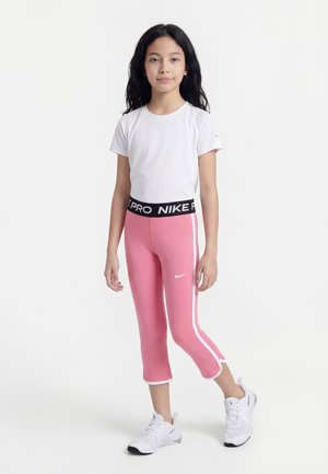 Fille debout portant un T-shirt blanc, un legging Nike Pro rose avec des bandes blanches, et des baskets blanches sur un fond uni.