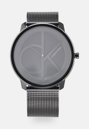 Montre avec bracelet en maille noire et cadran rond gris présentant le logo "CK" en relief et des aiguilles argentées. Design minimaliste avec des accents métalliques élégants.