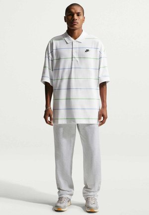 Uomo in piedi che indossa una polo bianca con sottili righe blu e verdi, pantaloni sportivi grigi e scarpe da ginnastica bianche su sfondo bianco.