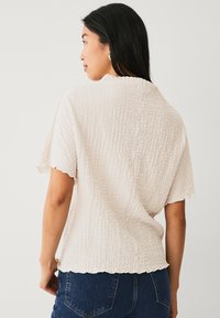Helle beige strukturiertes Oberteil mit kurzen Ärmeln, gewelltem Saum und lässigem Schnitt, kombiniert mit dunklen Jeans.
