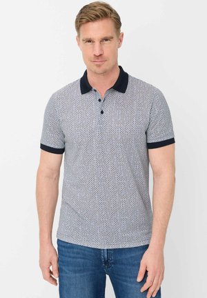 Blonder Mann, der ein kurzärmeliges Poloshirt mit blau-weißem Muster und blaue Jeans trägt, vor einem schlichten weißen Hintergrund steht.