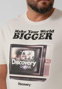Crèmegekleurde katoenen t-shirt met een retro televisie afbeelding en zwarte tekst die luidt: "Maak je wereld GROTER" en "Discovery Channel."