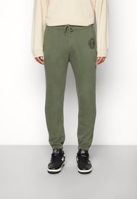 Olivgrüne Jogginghose aus weicher Baumwolle, mit Kordelzugbund und einem runden Logopatch am linken Oberschenkel.