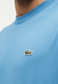 Άνδρας που φοράει ανοιχτό μπλε μπλουζάκι με στρογγυλή λαιμόκοψη, με ένα μικρό πράσινο λογότυπο κροκόδειλου κεντημένο στο στήθος.