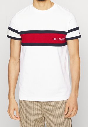 Mann trägt ein weißes kurzärmeliges T-Shirt mit marineblauen und roten horizontalen Streifen und dem Text "HILFIGER" auf der Brust.