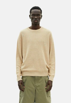 Beige strukturierter Pullover mit Rundhalsausschnitt und gerippten Details, getragen über olivgrünen Cargo-Hosen. Mit subtilen Abnutzungen an den Nähten.