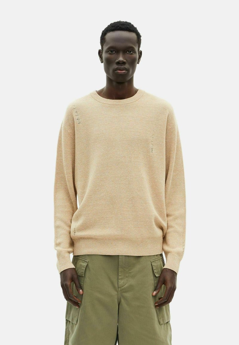 Beige strukturierter Pullover mit Rundhalsausschnitt und gerippten Details, getragen über olivgrünen Cargo-Hosen. Mit subtilen Abnutzungen an den Nähten.