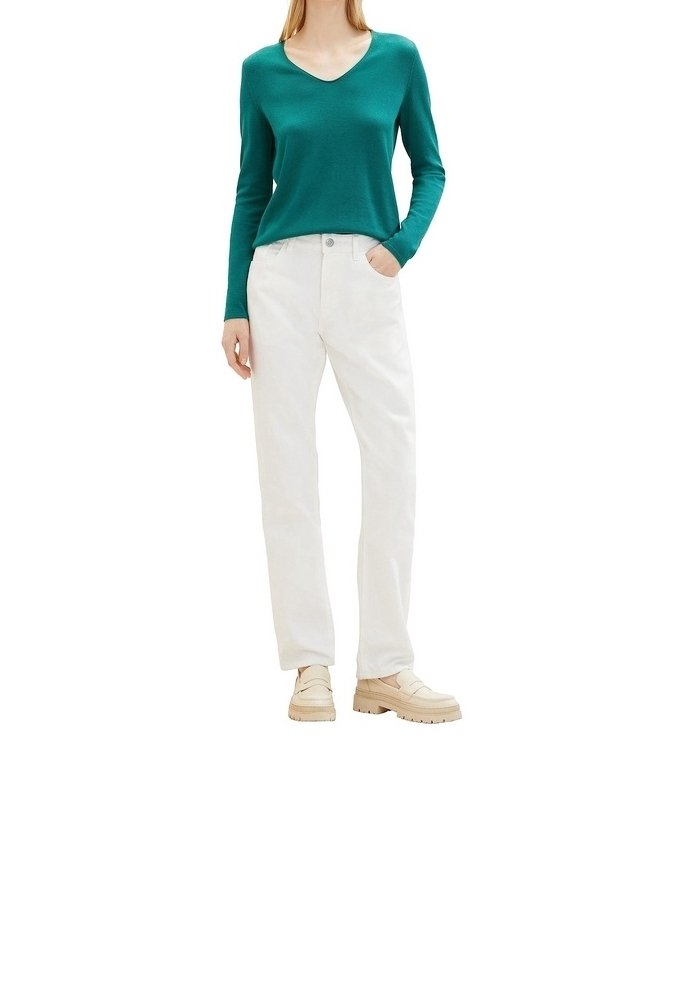 Femme portant un pull turquoise à manches longues et col en V, un pantalon blanc droit et des mocassins épais beige, debout avec une main dans la poche.