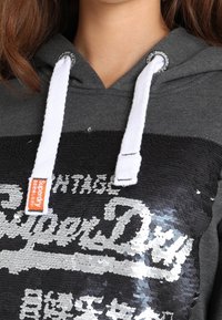 Felpa grigia con un logo ricamato in paillettes. Presenta cordini bianchi e un mix di paillettes opache e lucide in nero e argento.