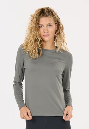 Femme aux cheveux blonds bouclés portant un t-shirt gris à manches longues, tenant l'ourlet, debout devant un fond blanc uni.
