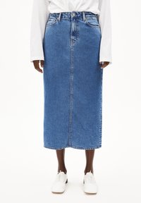 Falda de mezclilla en azul medio, con un corte recto, bolsillos frontales, trabillas para el cinturón y un dobladillo sin terminar, combinada con zapatos blancos de cordones.