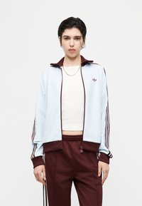 Νεαρό άτομο όρθιο, φορώντας ανοιχτό μπλε και μαροον αθλητική ζακέτα Adidas με αντίστοιχο παντελόνι, λευκό crop top και χρυσή αλυσίδα στον λαιμό.
