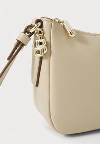 Sac à main en simili cuir beige de forme arrondie, avec des accessoires en or et des accents marron. Dispose d'une fermeture éclair et d'un détail en forme de charme avec le logo.