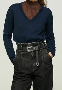 Marineblauer V-Ausschnitt-Pullover über einem braunen, gerippten Rollkragenpullover, kombiniert mit hoch taillierter schwarzer Denim-Hose. Enthält ein silbernes Gürtelschnallendetail.