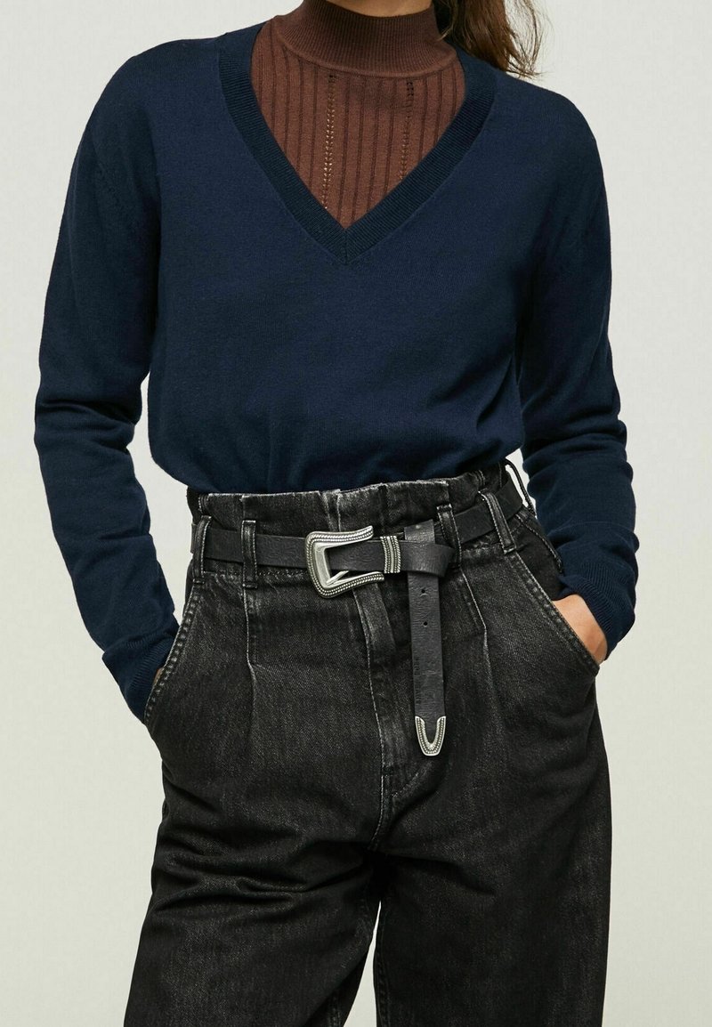 Marineblauer V-Ausschnitt-Pullover über einem braunen, gerippten Rollkragenpullover, kombiniert mit hoch taillierter schwarzer Denim-Hose. Enthält ein silbernes Gürtelschnallendetail.