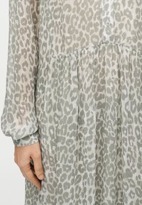 Ελαφρύ, διάφανο φόρεμα με γκρι και λευκό λεοπάρ μοτίβο. Διαθέτει κουμπιά στο μπροστινό μέρος, πλισέ φούστα και μακριά μανίκια.